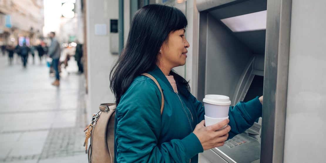 A person using an ATM machine inserting or withdrawing a card while holding cash. The person wears a green sweater and carries a small backpack with a beige strap.