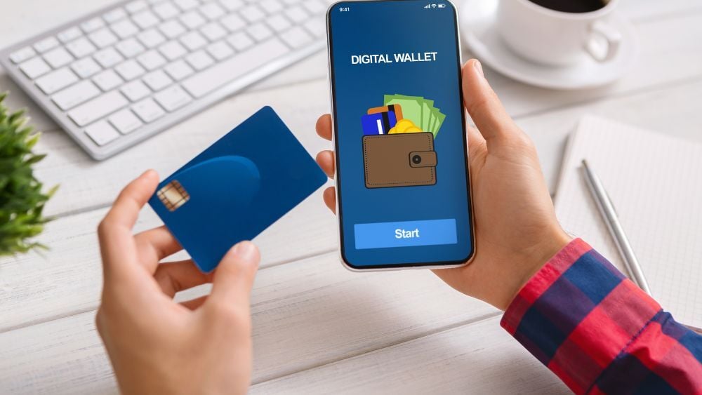 A person wearing a red and blue flannel, long sleeve shirt is holding a cell phone with their digital wallet app open in their right hand and a blue credit card in their left. There is a keyboard, coffee cup and saucer, pen, and notepad on the desk beneath them, along with a plant.