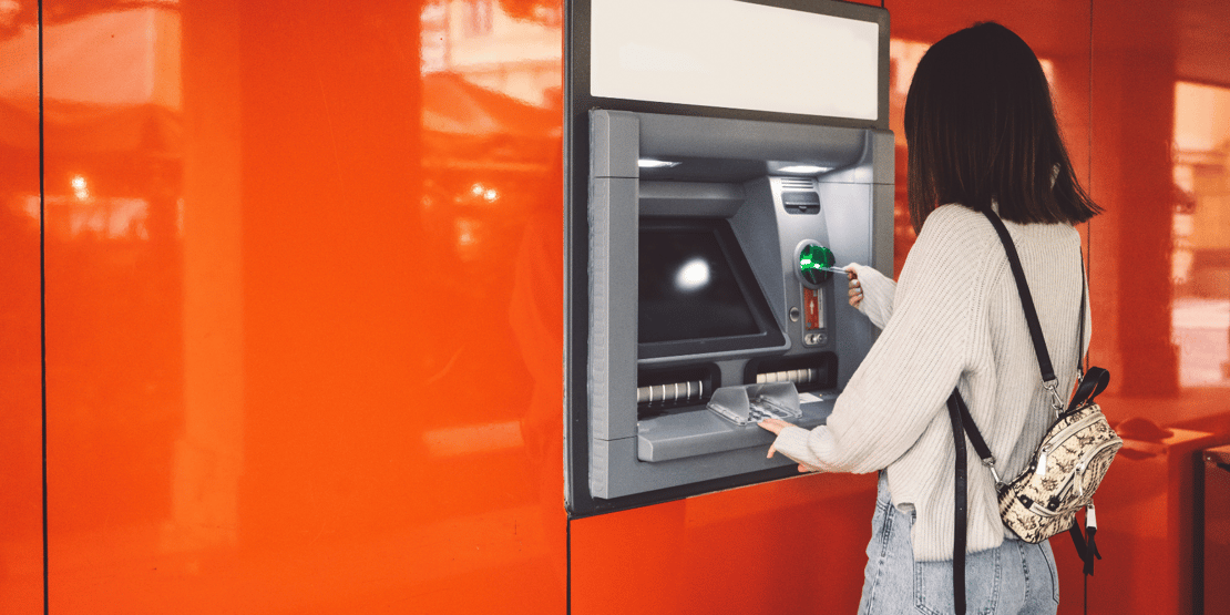 A person using an ATM machine attached to a bright orange wall, inserting or withdrawing a card while holding cash. The person wears a light sweater, blue jeans, and carries a small backpack with a black strap.
