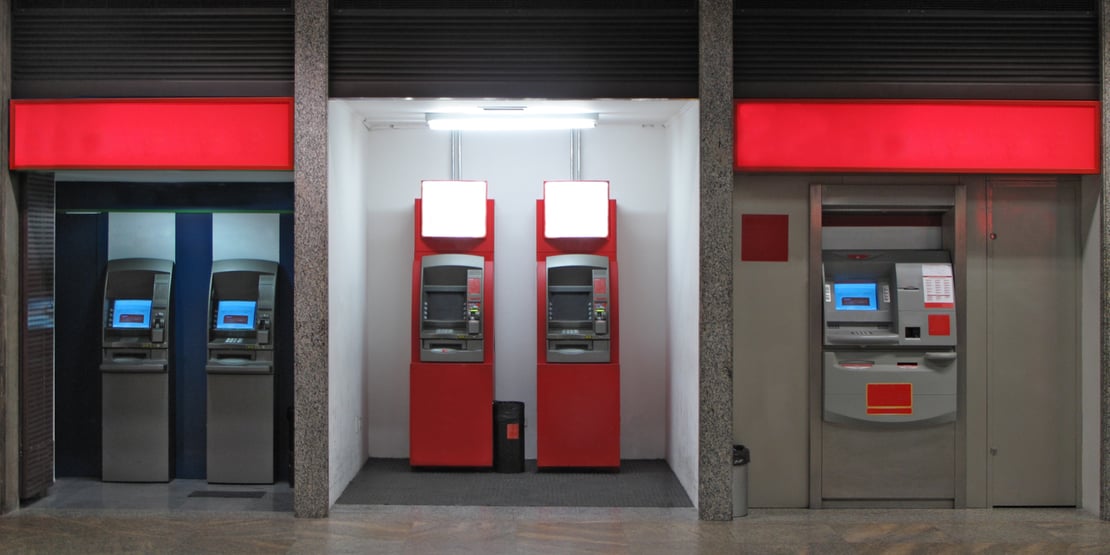 A row of ATMs from different banks, including OCBC, Maybank, UOB, and HSBC, in a brightly lit indoor area. A man with a backpack uses the OCBC ATM on the left, while a woman with a suitcase approaches the UOB ATM on the right.