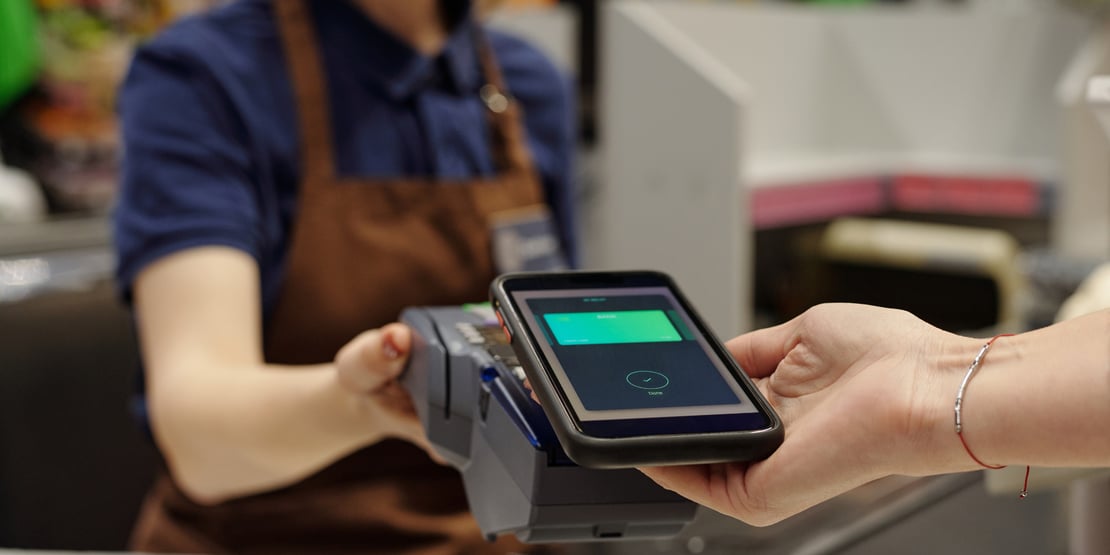 A person wearing a red and blue flannel, long sleeve shirt is holding a cell phone with their digital wallet app open in their right hand and a blue credit card in their left. There is a keyboard, coffee cup and saucer, pen, and notepad on the desk beneath them, along with a plant.