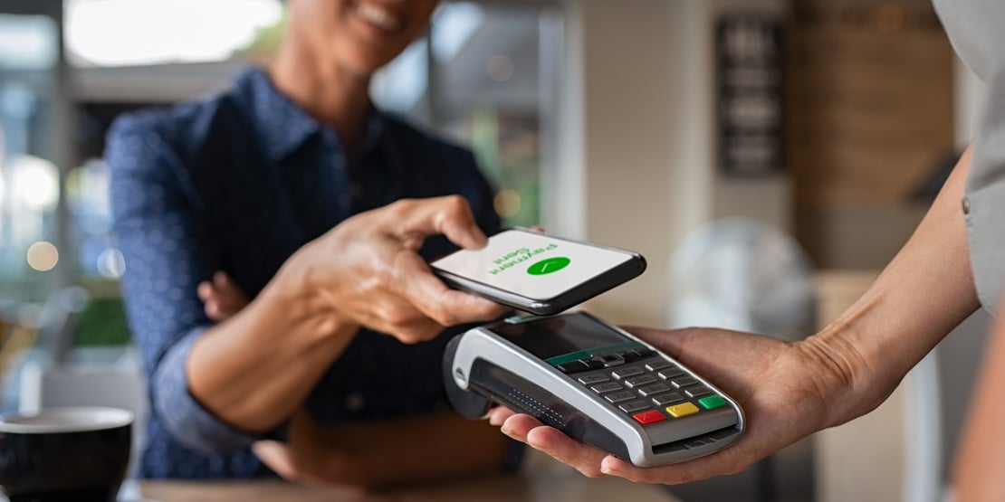 A smiling customer makes a contactless payment using a smartphone at a point-of-sale terminal in a modern café, illustrating the rise of digital and mobile payments.