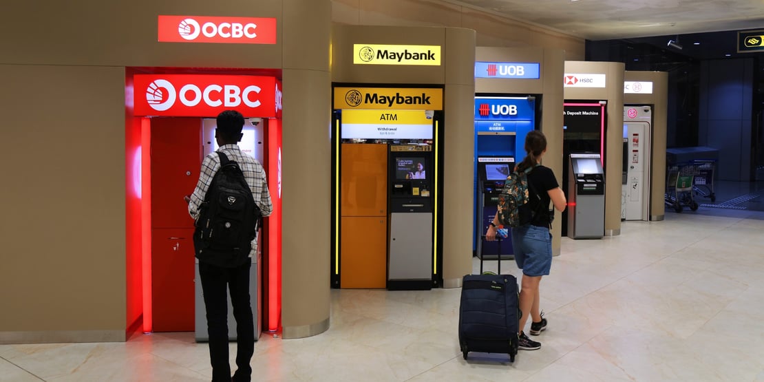 A row of ATMs from different banks, including OCBC, Maybank, UOB, and HSBC, in a brightly lit indoor area. A man with a backpack uses the OCBC ATM on the left, while a woman with a suitcase approaches the UOB ATM on the right.