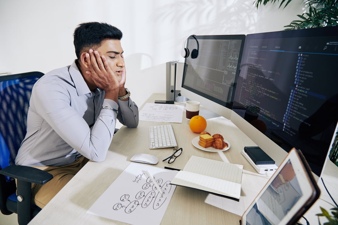A frustrated software developer sits at a cluttered desk with dual monitors displaying code. He rests his face in his hands, appearing stressed. The desk includes a keyboard, mouse, glasses, printed diagrams, a cup of coffee, an orange, and a plate of pastries.